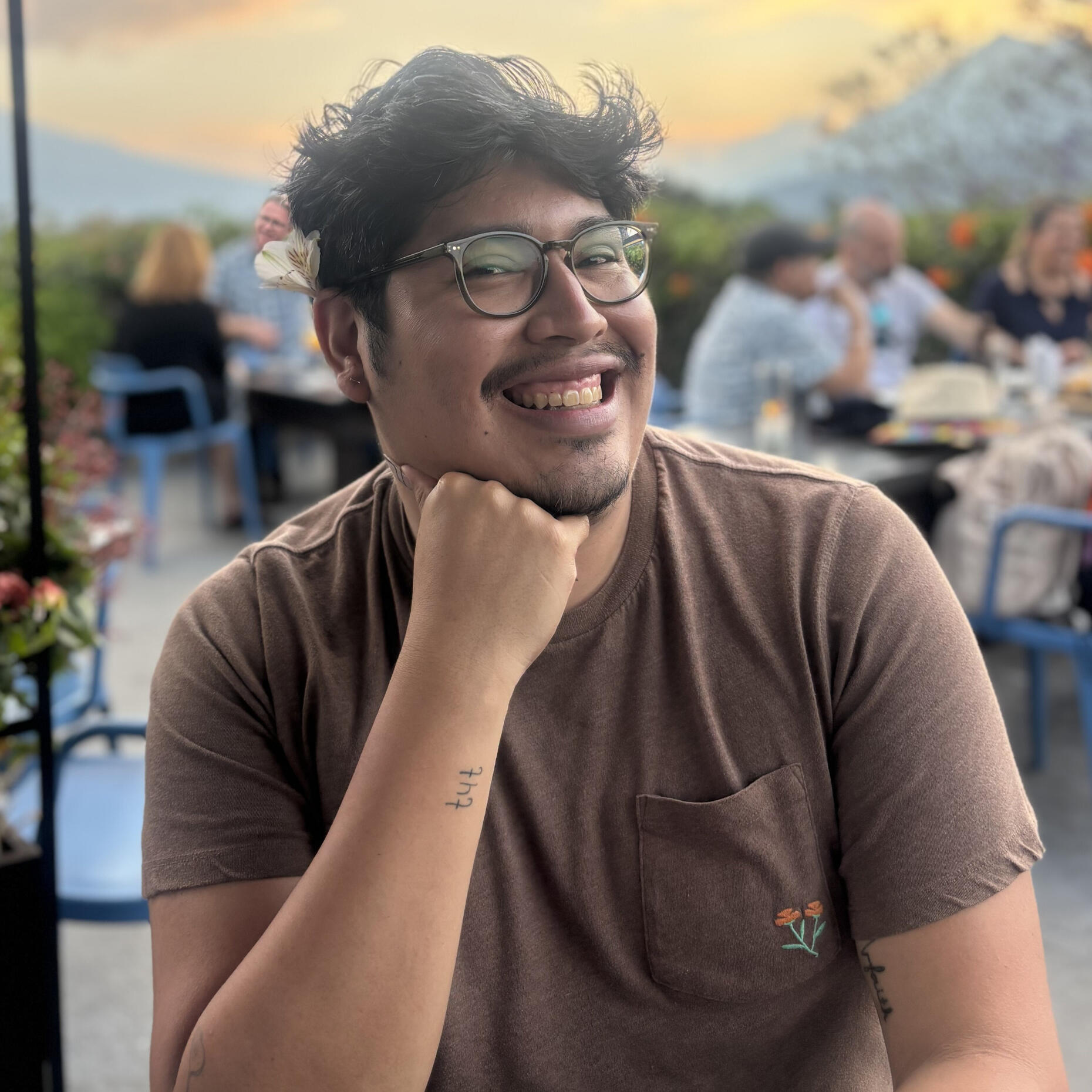 Joel smiles at the camera, posing with their right arm propped under their chin. Their head is turned slightly to the side, with a white flower tucked behind their right ear. They wear a brown t-shirt with a pocket, embroidered with orange poppies.