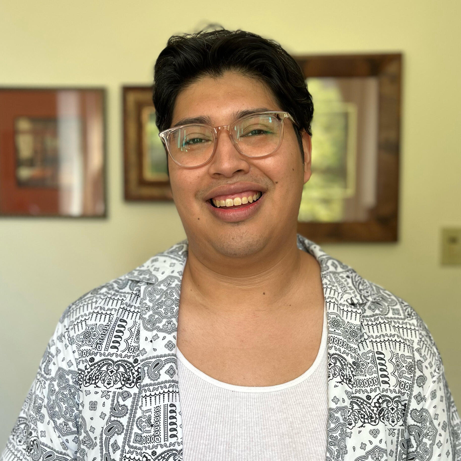 Joel stands against a beige backdrop with framed photos behind them. They smile at the camera, wearing a white tank top layered under a black-and-white bandana-patterned button-up shirt.