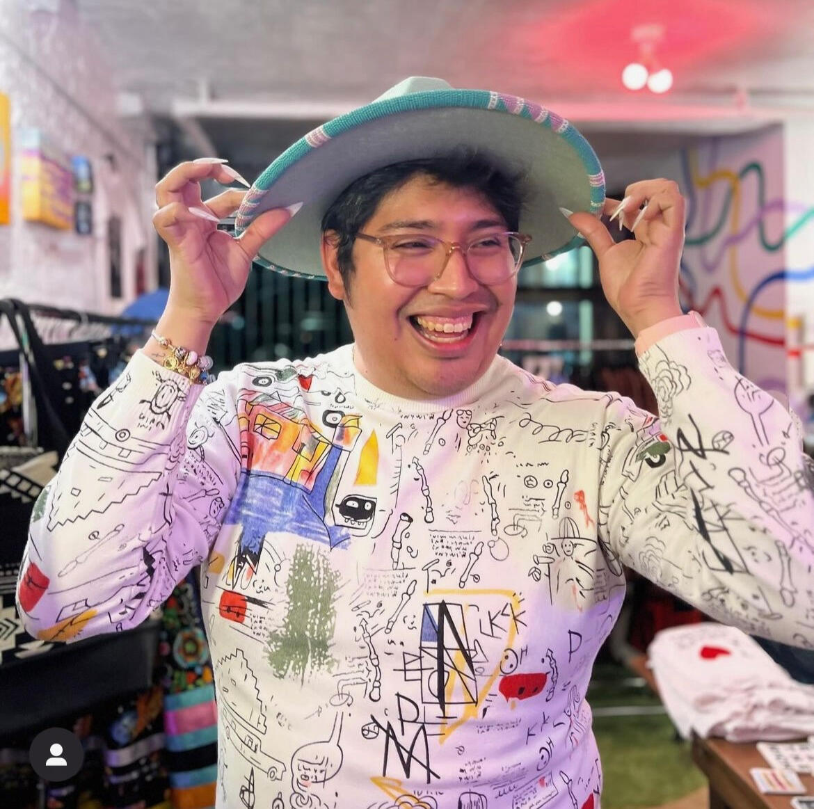 Joel is smiling and looking off-camera. They are wearing a white sweater with a Basquiat-inspired designs featuring various colors, including black, red, yellow, and blue. They hold a mint-colored felt hat with a beaded rim in green, mauve, and cream. Thei