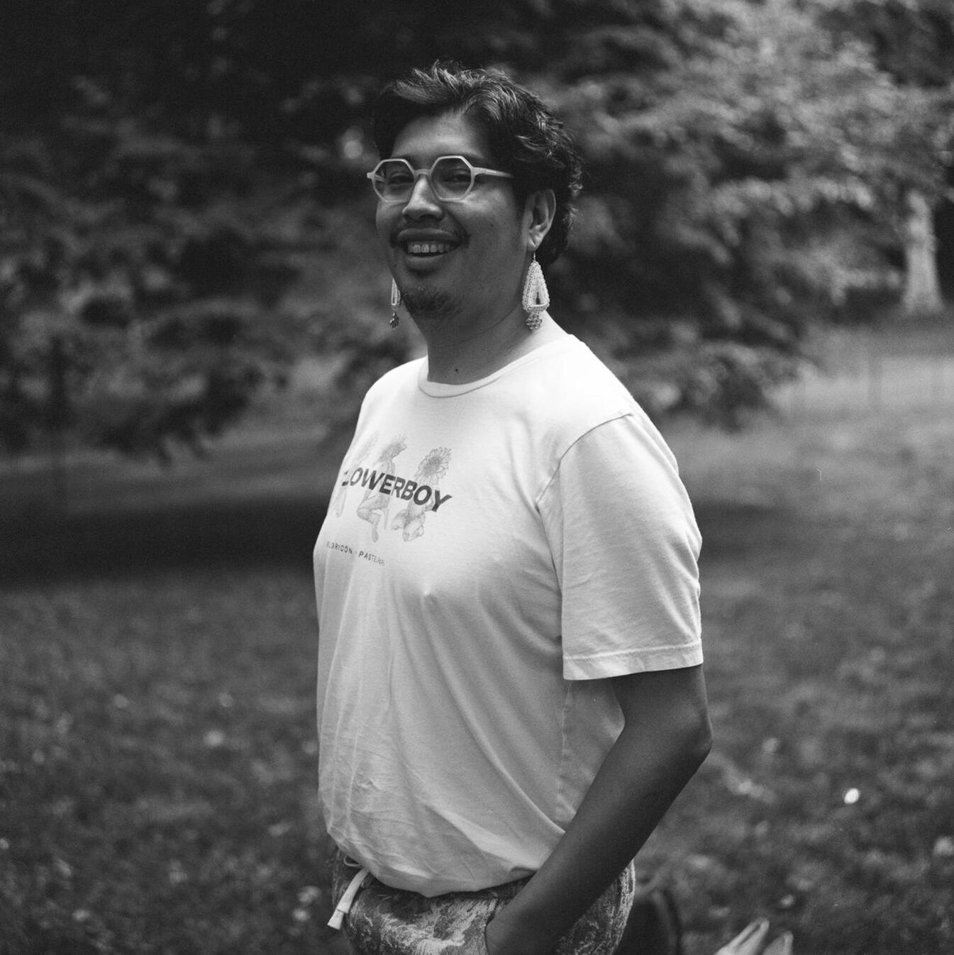 Joel is smiling at camera. They are in a 3/4 position, facing left. The photo is in black and white. Joel is wearing a Flowerboy shirt by Floricón. Their hands are in their pants pocket. A tree and greenery stand in the back.
