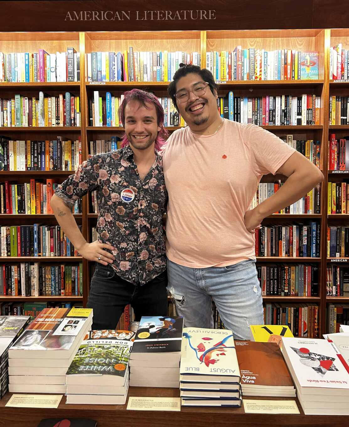 Ryan Cook (left) and Joel (right) pose together after a chance encounter at McNally Jackson. Both face the camera, each resting one hand on the other’s shoulder while their other arms wrap around each other’s waists. Joel wears a light orange shirt with a